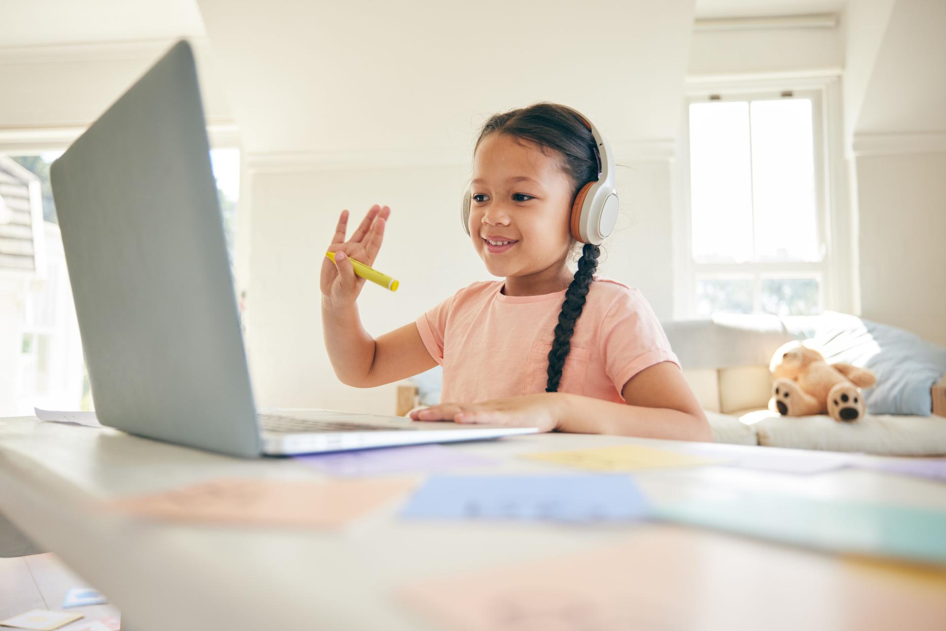 Laptop, elearning and child on a video call wave in a class doing homework in her bedroom at her house. Greeting, technology and kid student in a virtual online lesson for home school on a computer.